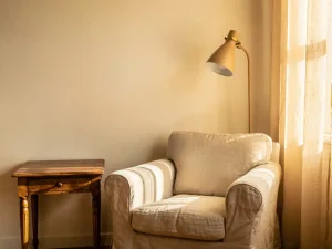 Vintage houten bijzettafel met linnen fauteuil en vloerlamp in terracotta woonkamer, warm middaglicht door sheer gordijnen.