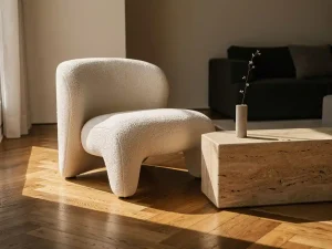 Sculpturele lounge chair in ivoor bouclé stof op eiken visgraatparket, naast een travertijnen bijzettafel met minimalistische vaas in zacht middaglicht.