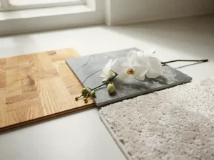 Drie vloermonsters van eiken parket, grijze steentegel en tapijt in warm zonlicht, met een witte orchideeëntak eroverheen.