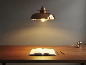 Designerhanglamp boven een eikenhouten bureau met open notitieboek, warm gouden licht contrasteert met koele grijze wanden.